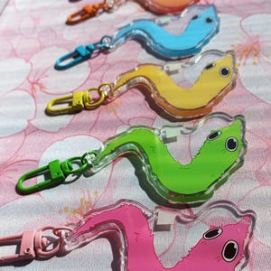 May include: A collection of six acrylic keychains shaped like fuzzy worms, each featuring a distinct colour including purple, orange, blue, yellow, green, and pink. Each keychain includes a matching coloured lobster clasp attached to the top.