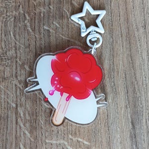 Könnte beinhalten: Ein klarer Acryl-Schlüsselanhänger mit einem roten Eis am Stiel-Design auf weißem Hintergrund. Das Eis am Stiel hat einen Holzstiel und tropft mit roter Flüssigkeit. Der Schlüsselanhänger hat einen weißen sternförmigen Verschluss.