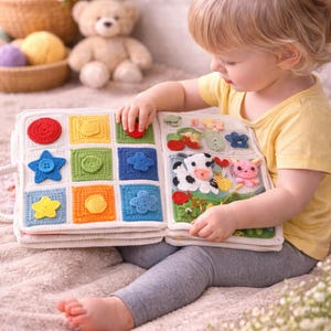 May include: A handmade crochet activity book for toddlers. The book features colorful squares with various shapes and textures, including a red circle, blue star, and yellow flower. The second page shows a cow, flowers, and a pig. The book is open, and a child is interacting with it.