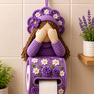 Könnte beinhalten: Ein handgefertigter, violetter Toilettenpapierhalter aus Häkelarbeit in Form einer Puppe mit floralem Design. Die Puppe hat braune Zöpfe und bedeckt ihre Augen. Der Halter ist mit weißen Gänseblümchen und lila Rosen verziert. Eine Rolle weißes Toilettenpapier ist sichtbar.