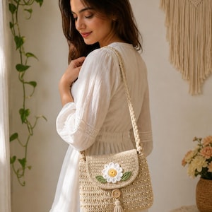 May include: A beige crochet crossbody bag with a daisy applique. The bag features a flap closure, a long strap, and a tassel. The daisy has white petals and a yellow center. The bag is approximately 9 inches wide and 7 inches tall.