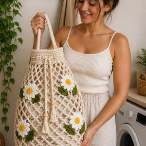 May include: Cream-colored crochet tote bag with a drawstring closure and daisy embellishments. The bag features a lattice design and is adorned with white and yellow flowers with green leaves. The handles are made of the same cream-colored yarn.