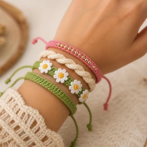 Puede incluir: Una colección de pulseras hechas a mano en varios estilos. Las pulseras incluyen una pulsera rosa con cuentas, una pulsera trenzada color crema, una pulsera de ganchillo verde con detalles de margaritas y una pulsera de ganchillo verde. Cada pulsera tiene un cierre ajustable.