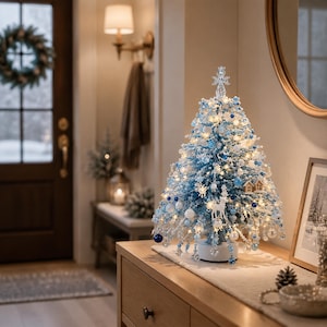 May include: A tabletop Christmas tree decorated with blue and white ornaments, including small spheres, snowflake shapes, and a white deer figurine. The tree is illuminated with warm white lights and topped with a clear, star-shaped ornament.