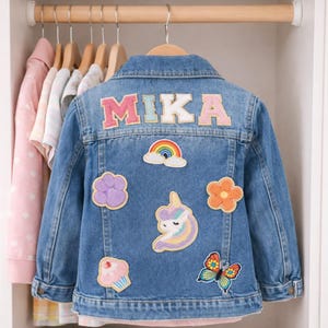 May include: A blue denim jacket with the name "MIKA" in colourful letters on the back. The jacket is decorated with patches of a rainbow, cloud, flower, cupcake, unicorn, and butterfly. The jacket is hanging on a wooden hanger.