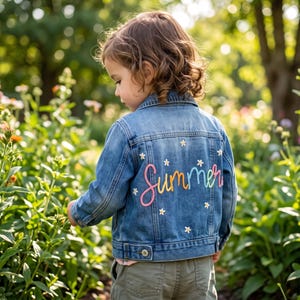 May include: A blue denim jacket featuring long sleeves, a collar, and a button-up front. The back of the jacket is decorated with the word Summer embroidered in a multi-coloured gradient thread, surrounded by small white embroidered flower motifs.