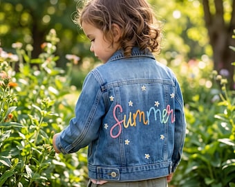 May include: A blue denim jacket featuring long sleeves, a collar, and a button-up front. The back of the jacket is decorated with the word Summer embroidered in a multi-coloured gradient thread, surrounded by small white embroidered flower motifs.