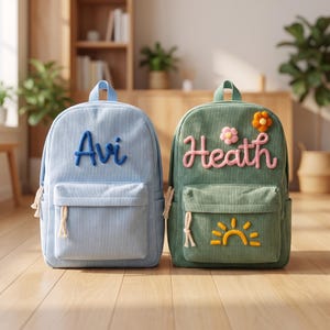 May include: Two corduroy backpacks, one light blue with the name Avi in blue yarn, and one sage green with the name Heath in pink yarn, a pink and orange flower, and a yellow sun motif on the front pocket, both featuring white rope zipper pulls.