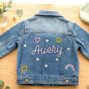 May include: A light blue denim jacket with the name "Avery" embroidered in pink script. The jacket features embroidered patches of a heart, smiley face, flowers, sun, and rainbow. The jacket is displayed on a wooden surface.