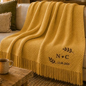 May include: A mustard yellow knit throw blanket with fringe trim, featuring a dark brown embroidered design of two leafy branches flanking the letters N and C, a small heart, and the date 12.08.2023 centred on the lower right corner.