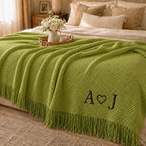 May include: A lime green textured knit throw blanket featuring a fringe trim along the bottom edge and the embroidered black letters A and J separated by a heart symbol. The blanket is draped over a bed with neutral-coloured pillows and bedding.