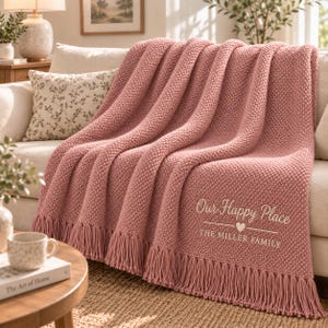 May include: A dusty rose knitted throw blanket with a textured bobble stitch pattern and long fringe trim along the bottom edge. The blanket features white embroidered text that reads Our Happy Place and THE MILLER FAMILY with a small heart icon.