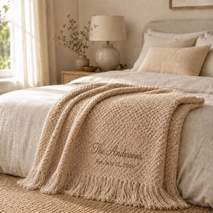 May include: A beige textured throw blanket with fringe trim, featuring dark brown embroidery that reads The Andersons and Est. July 12, 2026. The blanket is draped over a bed with matching beige bedding and pillows in a neutral-toned bedroom setting.