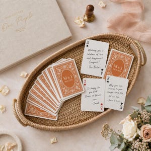 May include: A set of custom playing cards with terracotta-coloured backs featuring the initials M & J and the date 06.21.2026, displayed in a woven oval tray alongside cards with handwritten messages and a guest book labelled Our People.