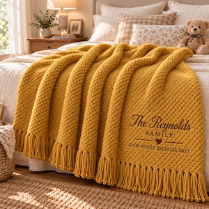Op de afbeelding: Een mosterdgele grofgebreide plaid met lange franjes aan de onderkant. De deken is voorzien van de geborduurde tekst The Reynolds FAMILY, een klein hartje en de zin GOOD PEOPLE BRIGHTER DAYS in donkerbruin.