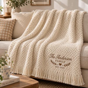 Peut inclure: Un jeté de canapé en tricot épais de couleur crème avec un motif texturé en relief et des franges. La couverture est brodée avec le texte The Anderson FAMILY, une petite icône de cœur et la date EST. 2020 en fil de couleur marron.