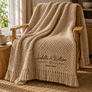 Op de afbeelding: Een beige, grofgebreide plaid met franjes, gedrapeerd over een houten stoel. De deken is voorzien van donkerbruin geborduurde tekst met Isabella & William, een klein hartje en de datum 2026.10.03 in het midden van de stof.