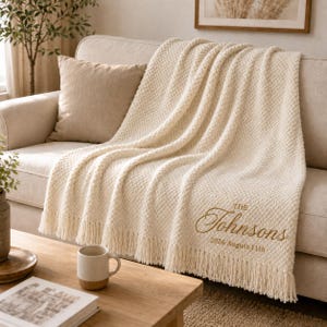 May include: A cream-coloured textured knit throw blanket with fringe trim, featuring embroidered gold text that reads "THE Johnsons" followed by the date "2026 August 11th" positioned in the lower right corner of the fabric.