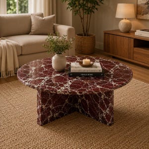 Puede incluir: Una mesa de centro redonda con un patrón de mármol rojo oscuro y blanco y una base de pedestal gruesa. Sobre la superficie hay un pequeño jarrón blanco con follaje verde, una vela blanca en un plato de madera y dos libros apilados titulados Live Beautifully y This Is Home.