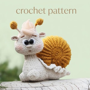 May include: A crocheted snail toy with a cream-colored body, a mustard-yellow shell, and large black eyes. The snail has small antennae with yellow pom-poms and a tuft of light pink hair. The text "crochet pattern" is above the snail.