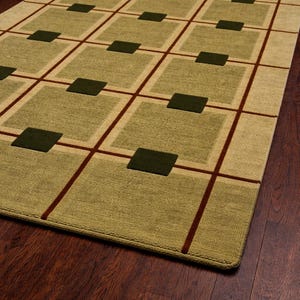 May include: A rectangular area rug with a geometric pattern. The rug features a grid design with olive green squares and dark brown lines on a light beige background. The rug is placed on a dark wood floor.