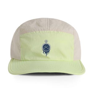 May include: A five-panel cap in cream and light green. The front panel features a navy blue pocket watch graphic with a dripping effect. The cap has a curved brim and a classic, sporty design.