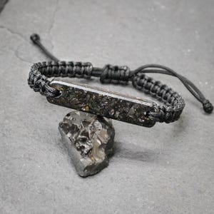 May include: A black cord bracelet featuring a rectangular centrepiece with a dark, textured, metallic-flecked surface. The bracelet is constructed with a woven macrame-style band and includes adjustable pull strings at the ends for a secure fit.