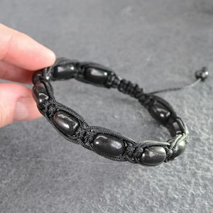 May include: A black bracelet featuring a series of smooth, oval-shaped black beads secured within a woven black cord pattern. The design includes an adjustable closure with two small black beads at the ends of the thin, trailing cord strings.