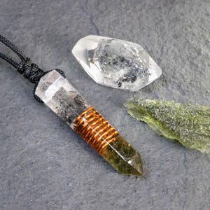 May include: A clear quartz crystal pendant with a copper wire wrap, on a black cord. Next to it are a clear quartz crystal and a piece of green moldavite, on a grey surface. The pendant is approximately 7.6 cm long.