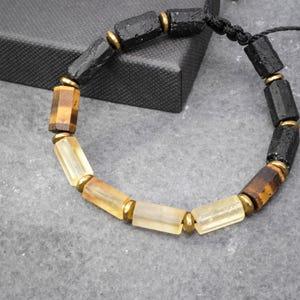 May include: A bracelet featuring cylindrical beads in black, brown tiger eye, and translucent yellow, separated by small metallic gold-toned spacers. The beads are strung on a black cord, creating a repeating pattern of dark and light earthy tones.