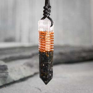 Op de afbeelding: Een heldere kristallen hanger met koperdraad en zwarte steeninsluitsels, opgehangen aan een zwart koord. De koperdraad is gewikkeld rond het middelste deel van het kristal, dat een puntige basis heeft.