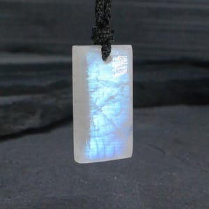 Puede incluir: Un colgante rectangular de piedra lunar con una superficie blanca translúcida y un efecto de adularescencia azul vibrante. La piedra está suspendida de un cordón negro fino que pasa por un pequeño orificio perforado en la parte superior central.