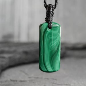 Op de afbeelding: Een groene malachiet hanger met een wervelend patroon, hangend aan een zwart koord. De rechthoekige steen is gepolijst en heeft een glanzende afwerking. Het koord is aan de bovenkant geknoopt, waardoor een lus ontstaat om als ketting te dragen.