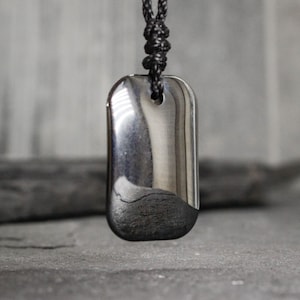 Peut inclure: Un pendentif rectangulaire avec une finition argentée polie et une moitié inférieure foncée et texturée. Le pendentif est suspendu à un cordon noir tressé. L'arrière-plan est un gris flou, avec une surface gris foncé en dessous.