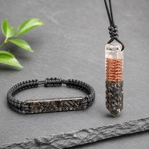 May include: A black cord bracelet featuring a rectangular bar filled with dark, textured mineral fragments and a matching pendant on a black cord, showcasing a clear top section, a coiled copper wire middle, and a dark mineral base.