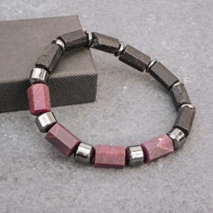 Può includere: Un braccialetto di perline con cilindri alternati di pietra nera dalla consistenza ruvida e pietra rosa scuro liscia e sfaccettata, separate da piccole perline distanziatrici metalliche color argento lucido in un motivo circolare.