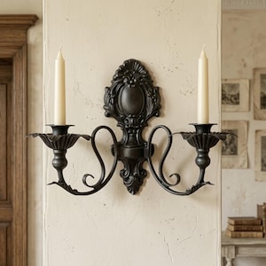 May include: A black wall-mounted candle holder featuring two arms with scalloped bobeches and a central decorative backplate with ornate scrollwork. Two off-white taper candles are inserted into the holders, creating a symmetrical design.