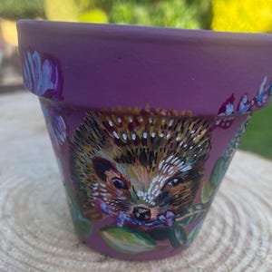 May include: A hand-painted, purple ceramic flower pot with a detailed hedgehog illustration. The hedgehog has brown and white quills, a detailed face, and floral accents. The pot is sitting on a wooden surface.