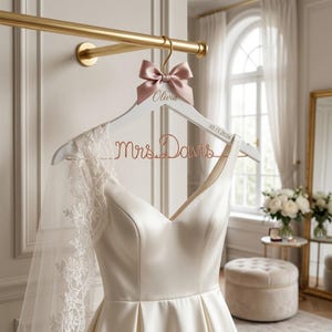 Puede incluir: Una percha blanca de madera para vestido de novia con un lazo rosa y el nombre "Olivia" en escritura dorada. La percha presenta las palabras "Mrs. Davis" en alambre de cobre y la fecha "10.15.2026". Un vestido de novia de satén blanco cuelga de la percha.