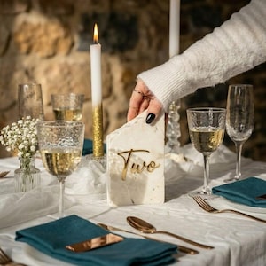 May include: A table setting with a white tablecloth, teal napkins, and gold-colored cutlery. A marble table number with the word "Two" in gold is displayed. Lit candles and glasses of wine complete the elegant arrangement.