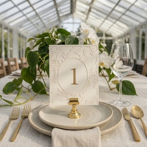 Könnte beinhalten: Tischdekoration für eine Hochzeit mit einer cremefarbenen Tischnummerkarte mit der goldenen Zahl 1. Die Karte wird von einem goldenen Clip gehalten und liegt auf einem weißen Teller. Goldenes Besteck und ein Weinglas sind ebenfalls vorhanden.
