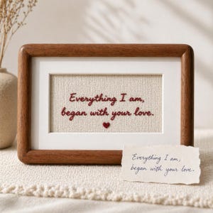 May include: A framed piece of art featuring the embroidered words "Everything I am, began with your love." The text is in red thread on a beige fabric, with a small red heart. The frame is made of dark wood.