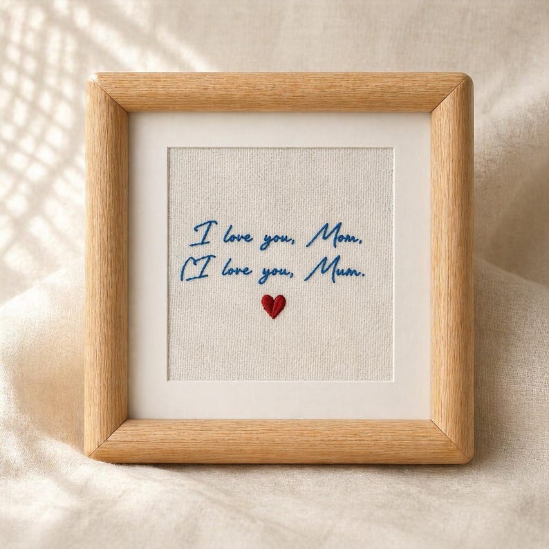 May include: A framed embroidery with the words "I love you, Mom" in blue thread, and a red heart. The frame is made of light-colored wood, and the background is a neutral fabric.