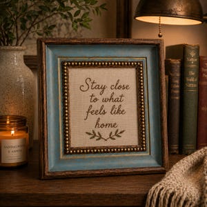 May include: A framed artwork with the text "Stay close to what feels like home." The frame has a distressed blue and brown finish with a beaded inner border. A lit candle and books are in the background, creating a cozy, homey atmosphere.