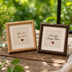 May include: Two framed artworks with heartfelt messages. One reads "You are my home" with a red heart, the other says "Love you always, Mom" with a red heart. The frames are wood, one light and one dark.