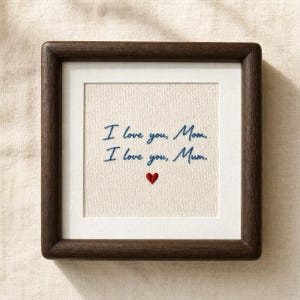 May include: A framed embroidered artwork with the text "I love you, Mom. I love you, Mum." in blue script. A small red heart is embroidered below the text. The frame is dark brown.