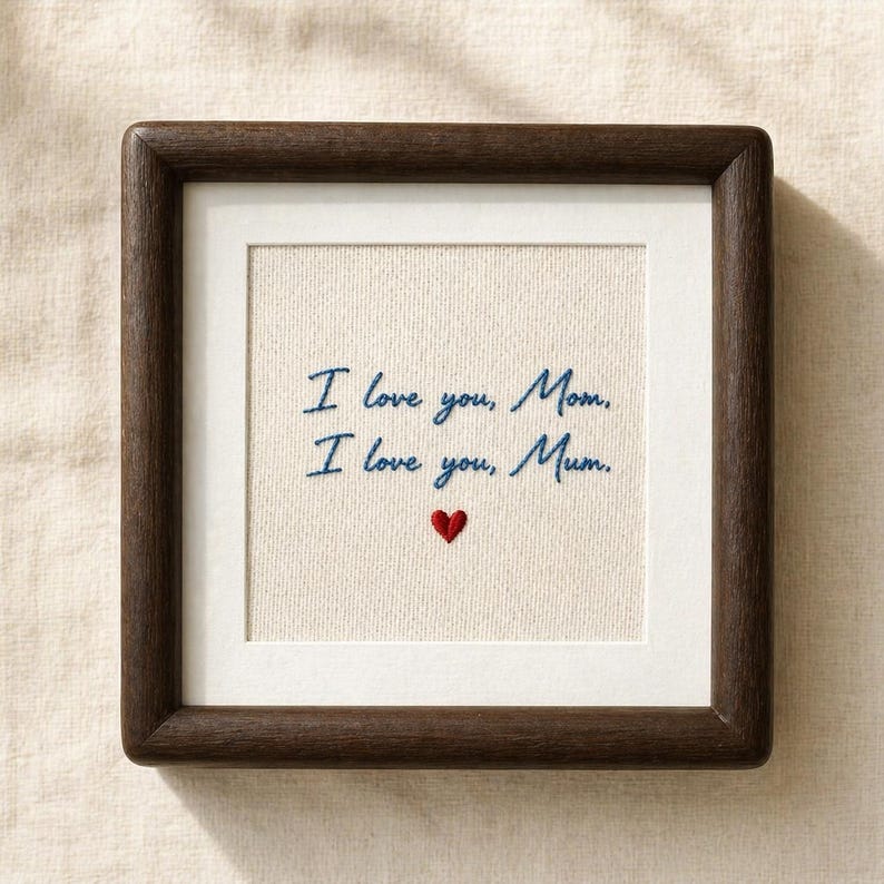 May include: A framed embroidered artwork with the text "I love you, Mom. I love you, Mum." in blue script. A small red heart is embroidered below the text. The frame is dark brown.