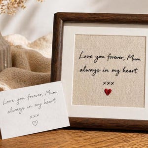 May include: A framed artwork with the text "Love you forever, Mum always in my heart" embroidered on a beige fabric. A small red heart is stitched below the text. A handwritten card with the same text and a heart is next to the frame.