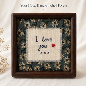 May include: A square dark wood frame containing a piece of fabric with a dark teal background and a repeating cream daisy pattern. Centered is a cream fabric square with frayed edges featuring the hand-stitched text I love you xxx and a red heart.