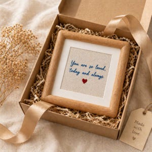 May include: A framed artwork with the text "You are so loved, today and always" and a red heart, presented in a gift box with ribbon and a tag that says "Made with love". The frame is wood.